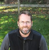 man in black vest and gray shirt is smiling at the camera with green grass and leaves in the background